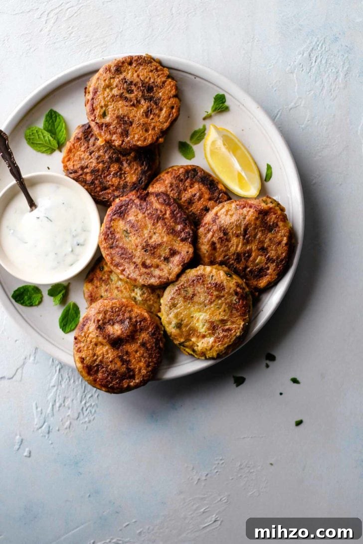 A white plate with Shami Kebab alongside a bowl of raita, lemon wedge and garnished with mint leaves.