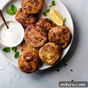 A white plate with Shami Kebab alongside a bowl of raita, lemon wedge and garnished with mint leaves.