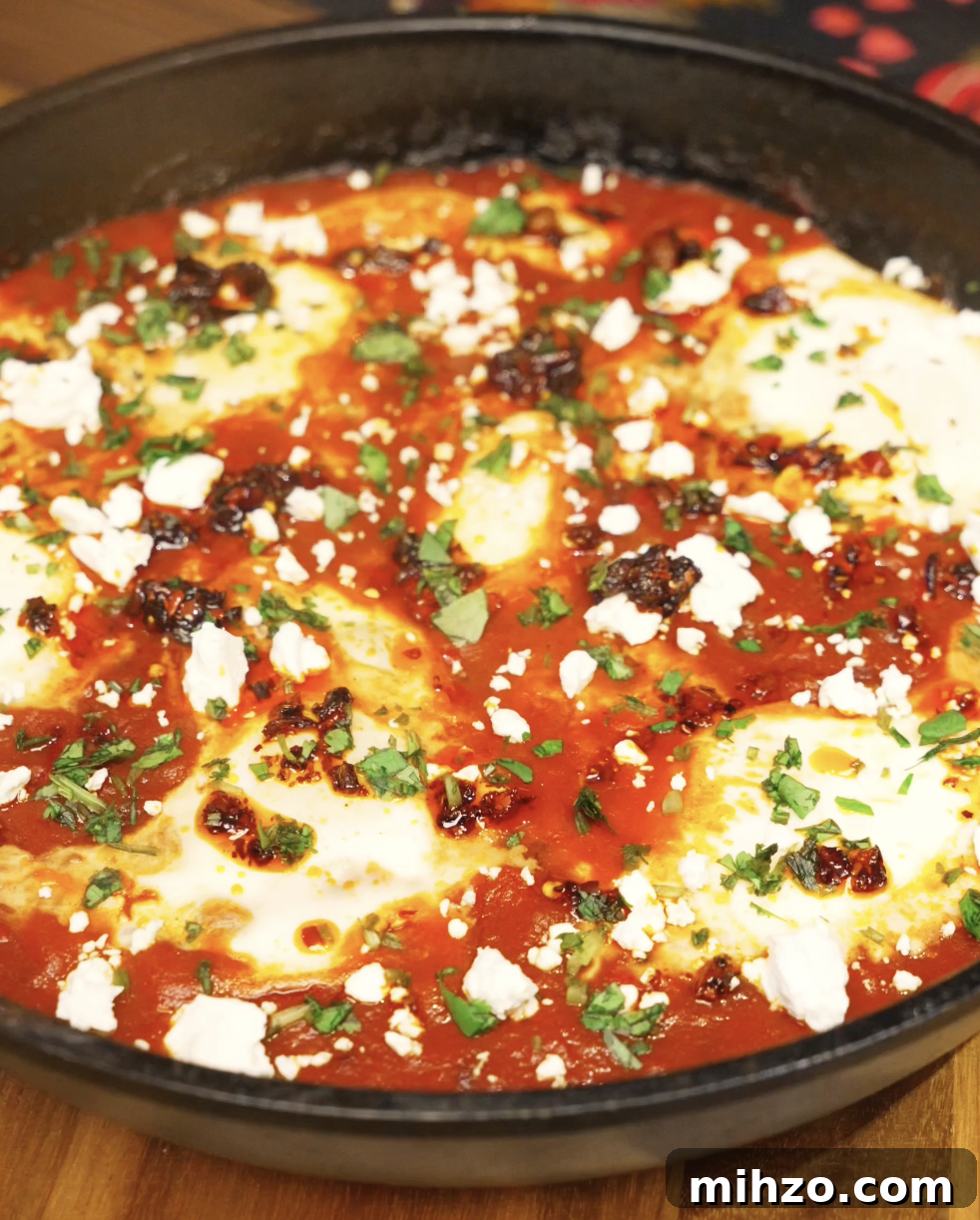 Chili oil shakshuka simmering in a skillet with gently poached eggs set in a rich tomato sauce, finished with feta, herbs, and chili oil.