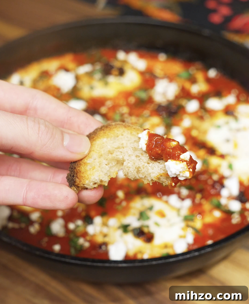 A piece of toasted bread dipped into chili oil shakshuka, scooping up tomato sauce, soft egg, and crumbled feta for a perfect bite.