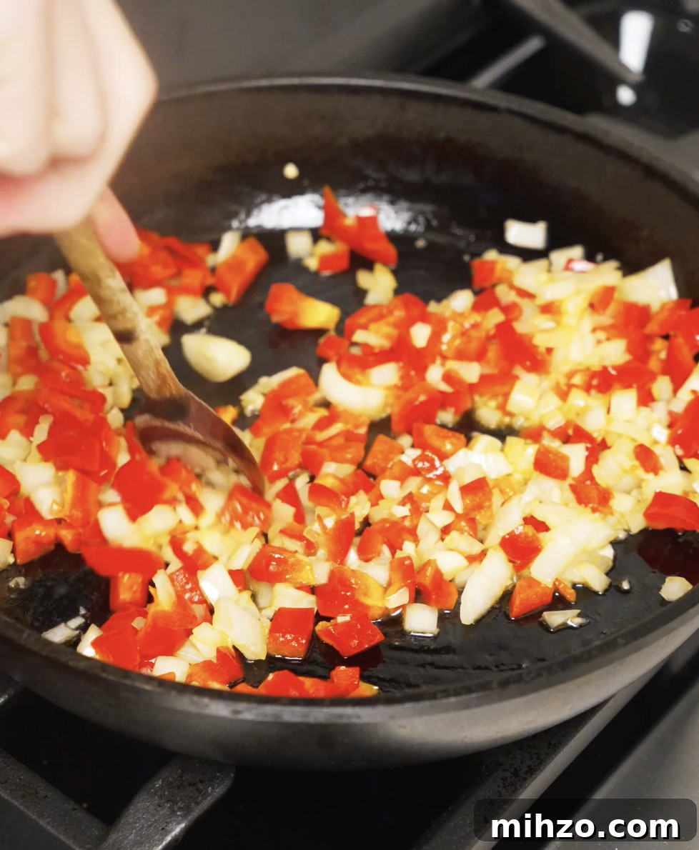 Diced red bell peppers and onions being sautéed in oil until softened and fragrant in a skillet.