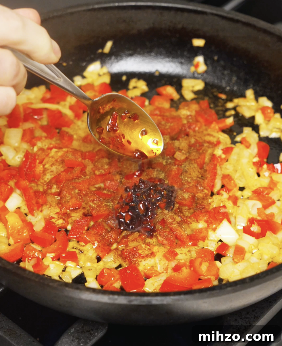 Chili oil and spices being spooned into sautéed peppers and onions, blooming the aromatics for the shakshuka sauce.