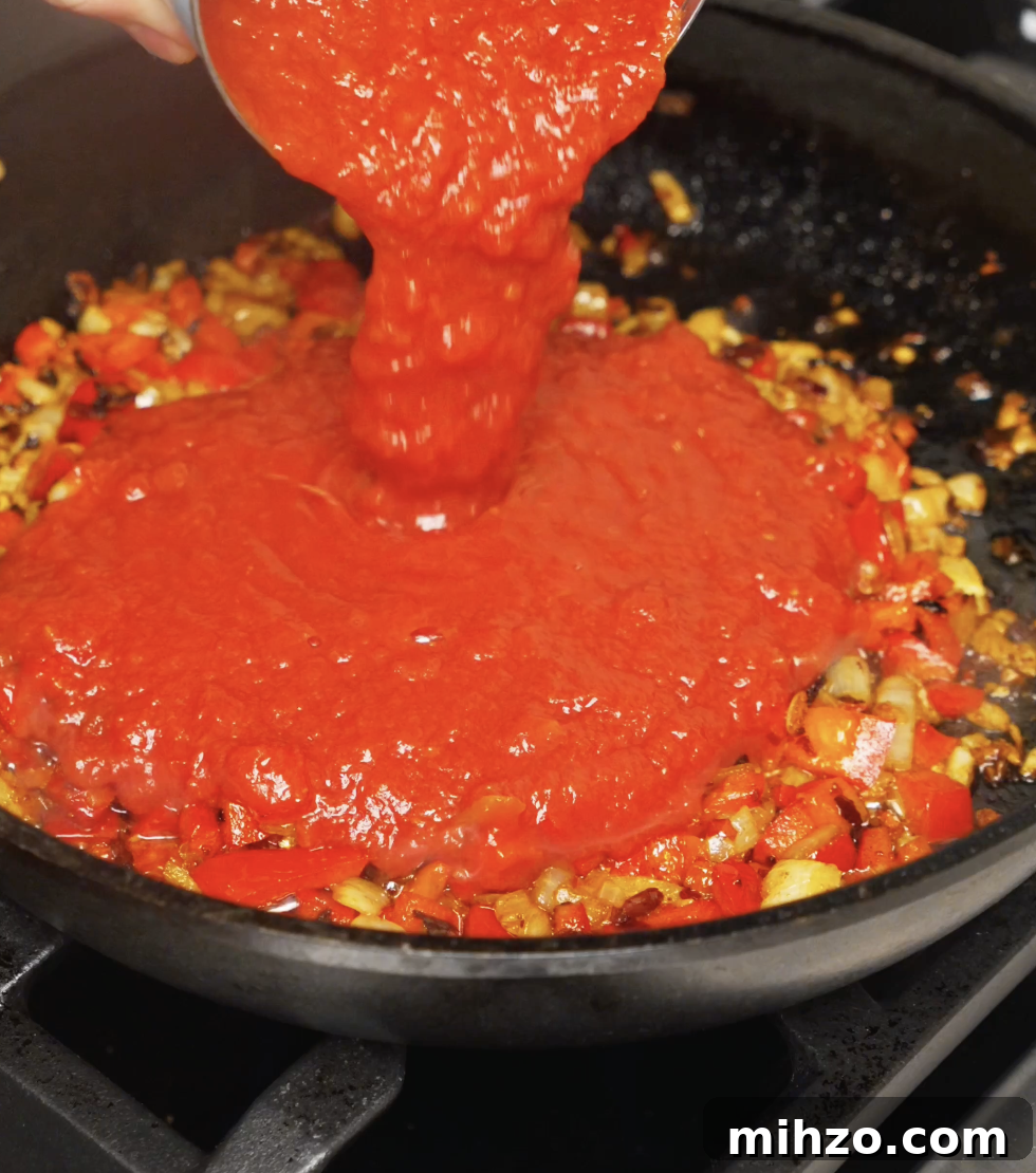 Crushed tomatoes being poured into a pan of sautéed onions, red bell peppers, garlic, and spices, forming the base for chili oil shakshuka.