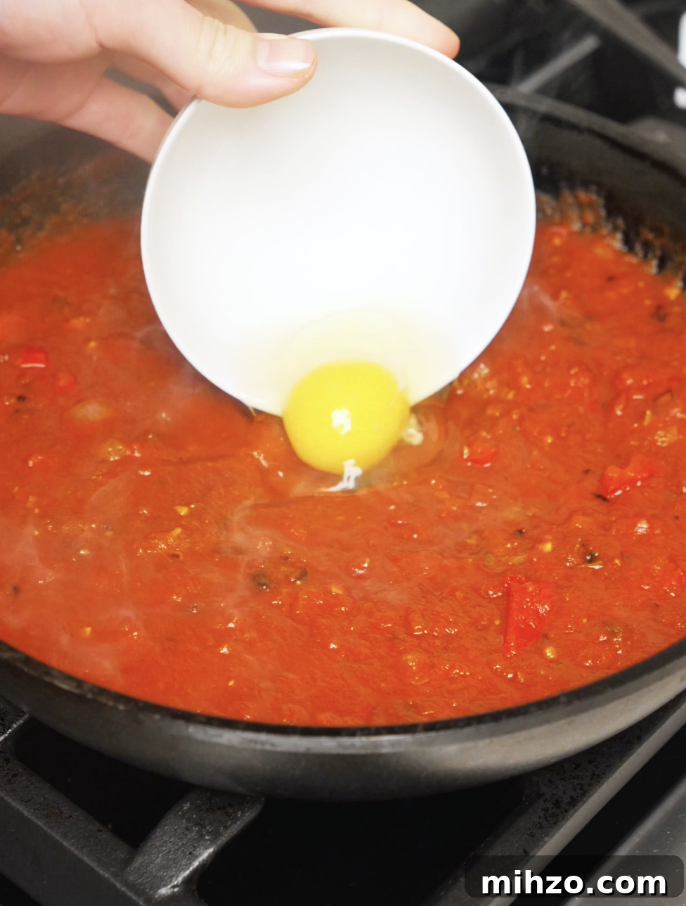 An egg being carefully cracked into a well in the simmering chili oil tomato sauce to poach directly in the shakshuka.