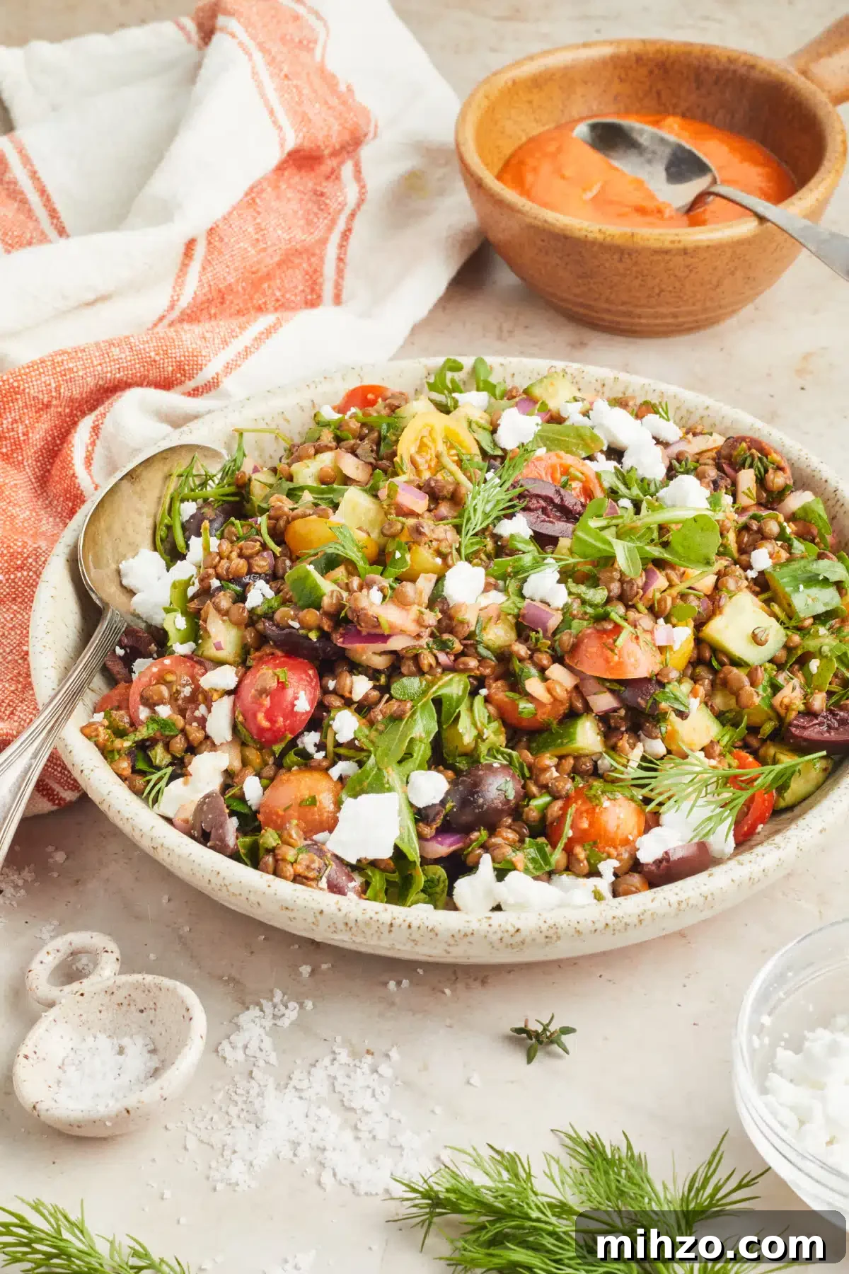 Greek Lentil Salad @spabettie #glutenfree #vegan #dairyfree #greeksalad #lentilsalad #dinner #recipe Serving bowl of colorful Mediterranean lentil salad consisting of lentils, sliced cherry tomatoes, sliced cucumber, fresh herbs, dairy free feta cheese. A small bowl of bright orange sun dried tomato dressing sits on the side with an orange and white striped cloth napkin.