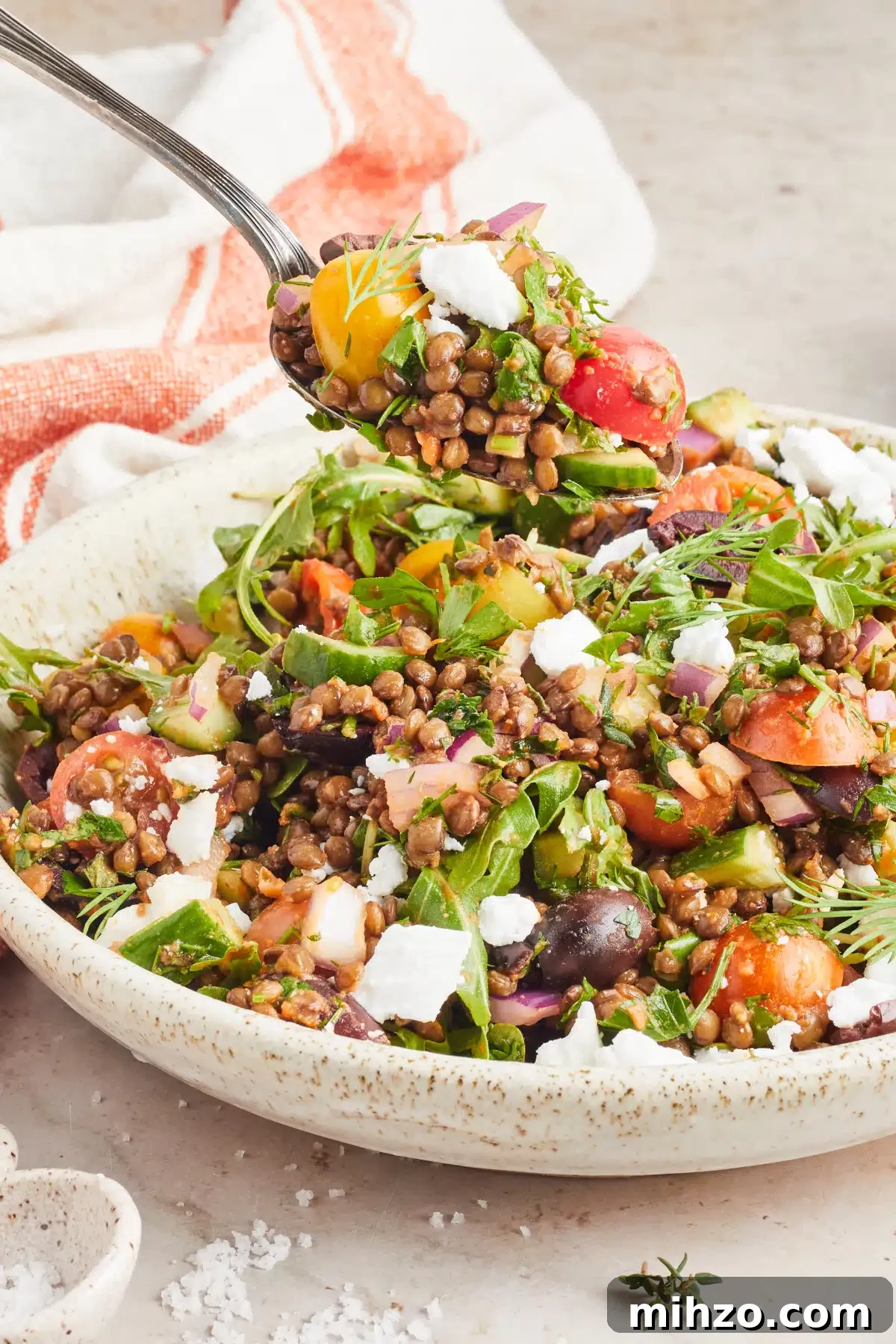 Mediterranean Salad @spabettie #glutenfree #vegan #dairyfree #greeksalad #lentilsalad #dinner #recipe A spoon of salad hovers over a serving bowl of colorful Greek salad with lentils, sliced cherry tomatoes, sliced cucumber, fresh herbs, dairy free feta cheese. A small bowl of bright orange sun dried tomato dressing sits on the side with an orange and white striped cloth napkin.