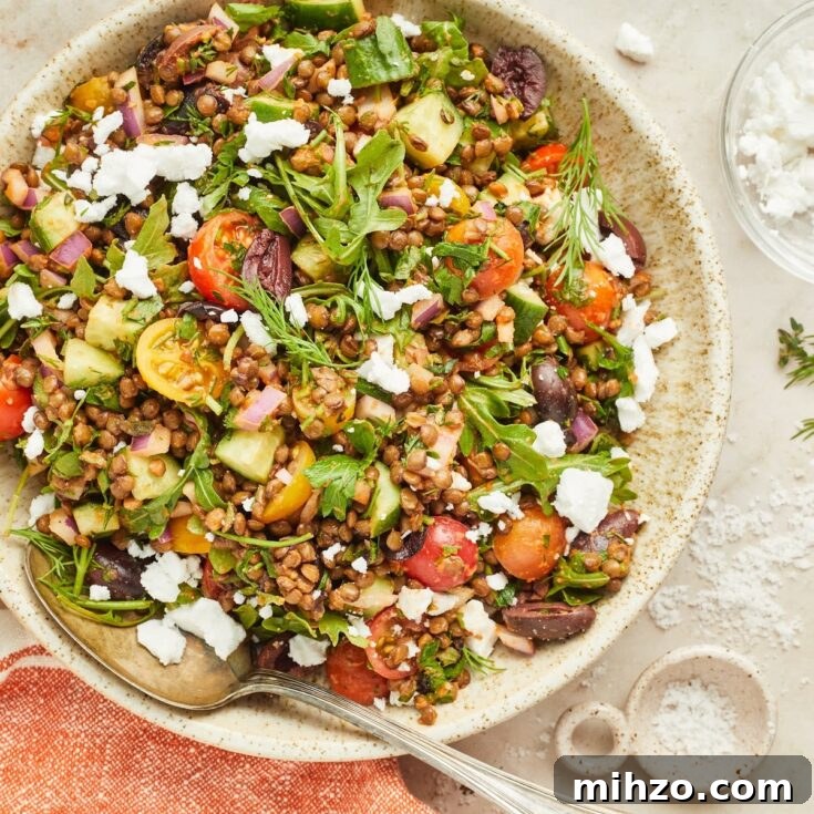 Overhead view of a serving bowl of colorful Greek lentil salad consisting of lentils, sliced cherry tomatoes, sliced cucumber, fresh herbs, dairy free feta cheese.
