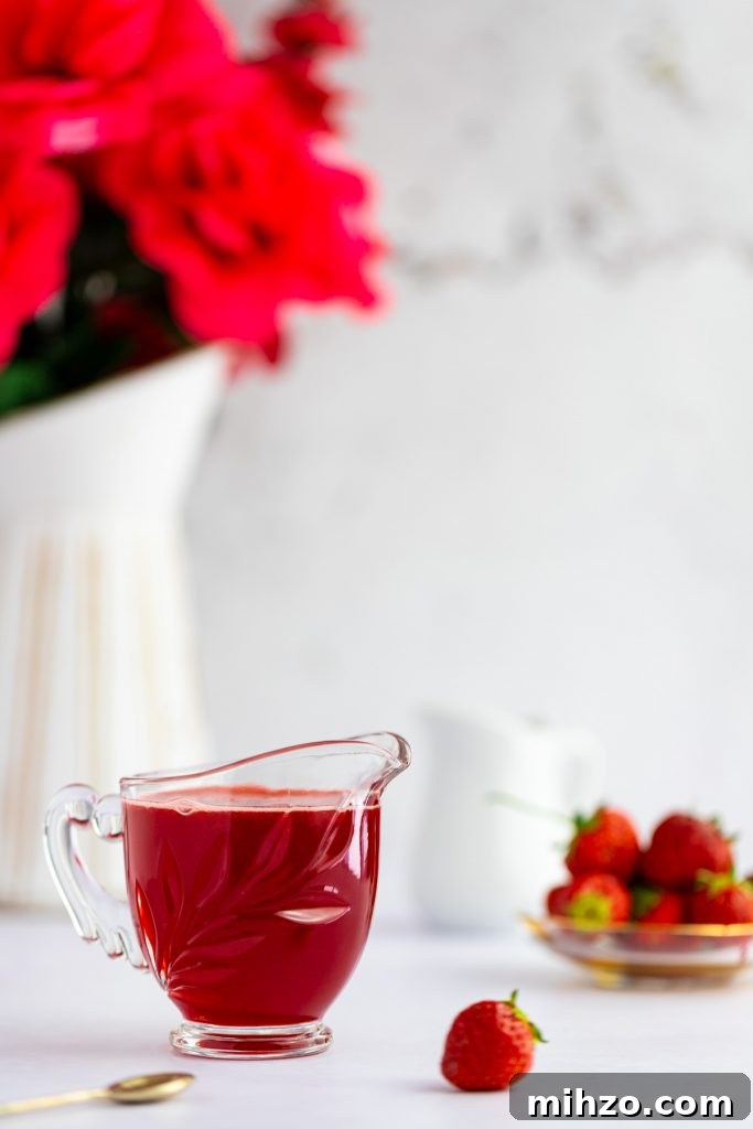 Strawberry Simple Syrup Recipe for Drinks and Desserts 6 A small glass container filled with red strawberry simple syrup, a pile of strawberries in the background and a small gold spoon in the foreground.