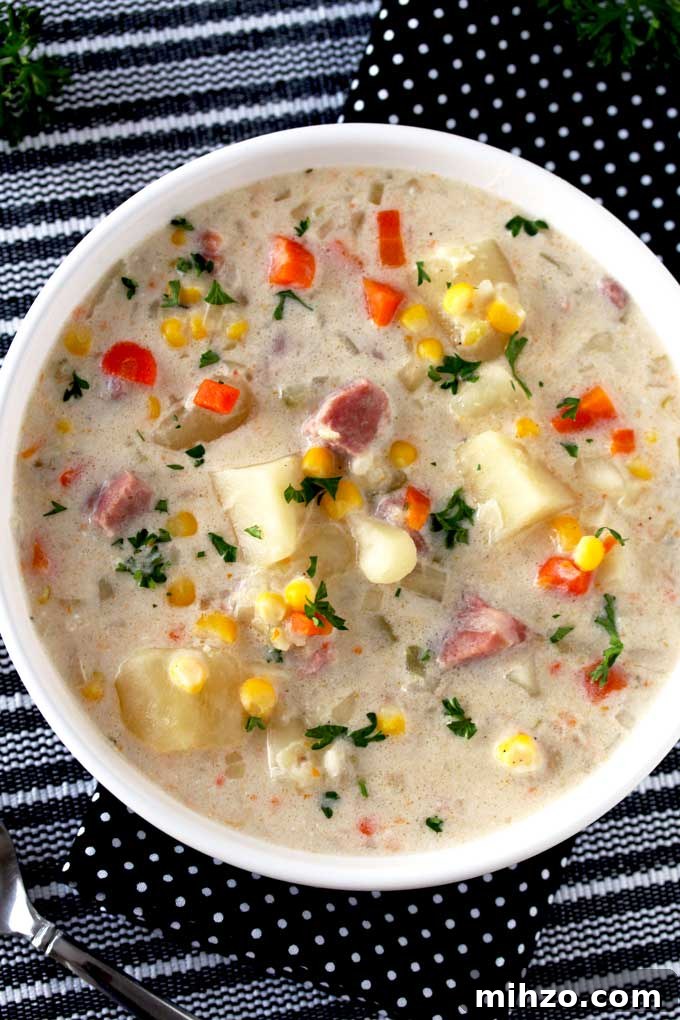 Slow Cooker Creamy Ham and Potato Soup Recipe 4 Top view of a white bowl filled with creamy ham and potato soup with potatoes, corn and carrots and garnished with chopped parsley sitting on a striped black and white table mat.
