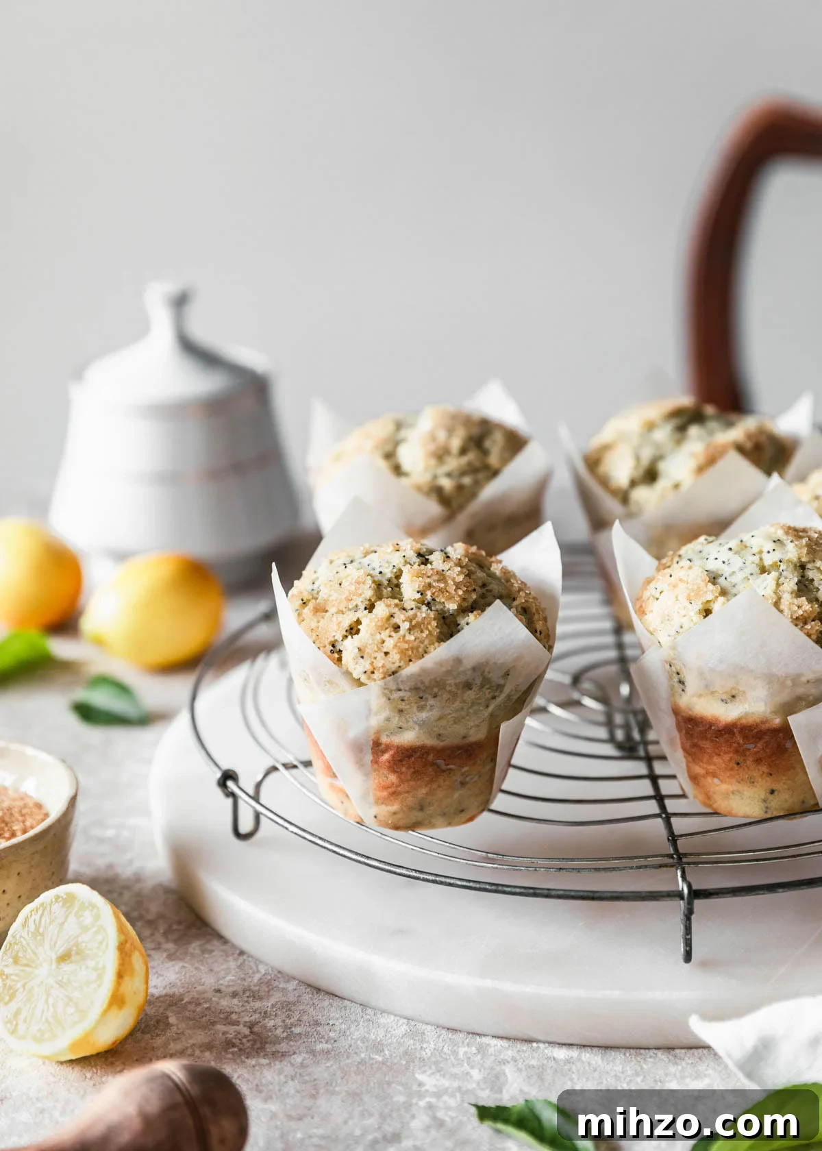 Ricotta Lemon Poppy Seed Muffins with Zesty Citrus Glaze 2 Ricotta lemon poppy seed muffins on a circular cooling rack next to lemons and a white sugar bowl on a beige speckled counter.