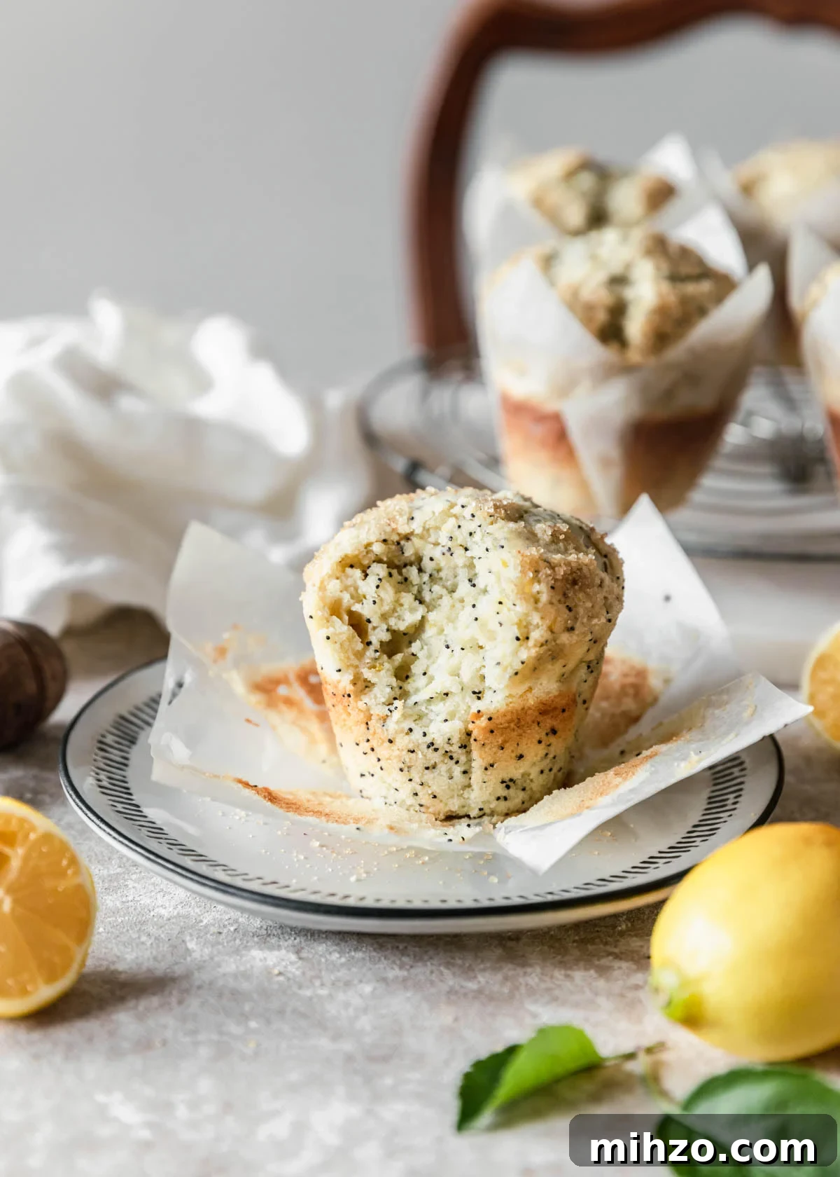 Ricotta Lemon Poppy Seed Muffins with Zesty Citrus Glaze 15 A close shot of a ricotta lemon poppy seed muffin with a bite taken out of it on a white plate with more muffins and lemons in the background on a beige table.