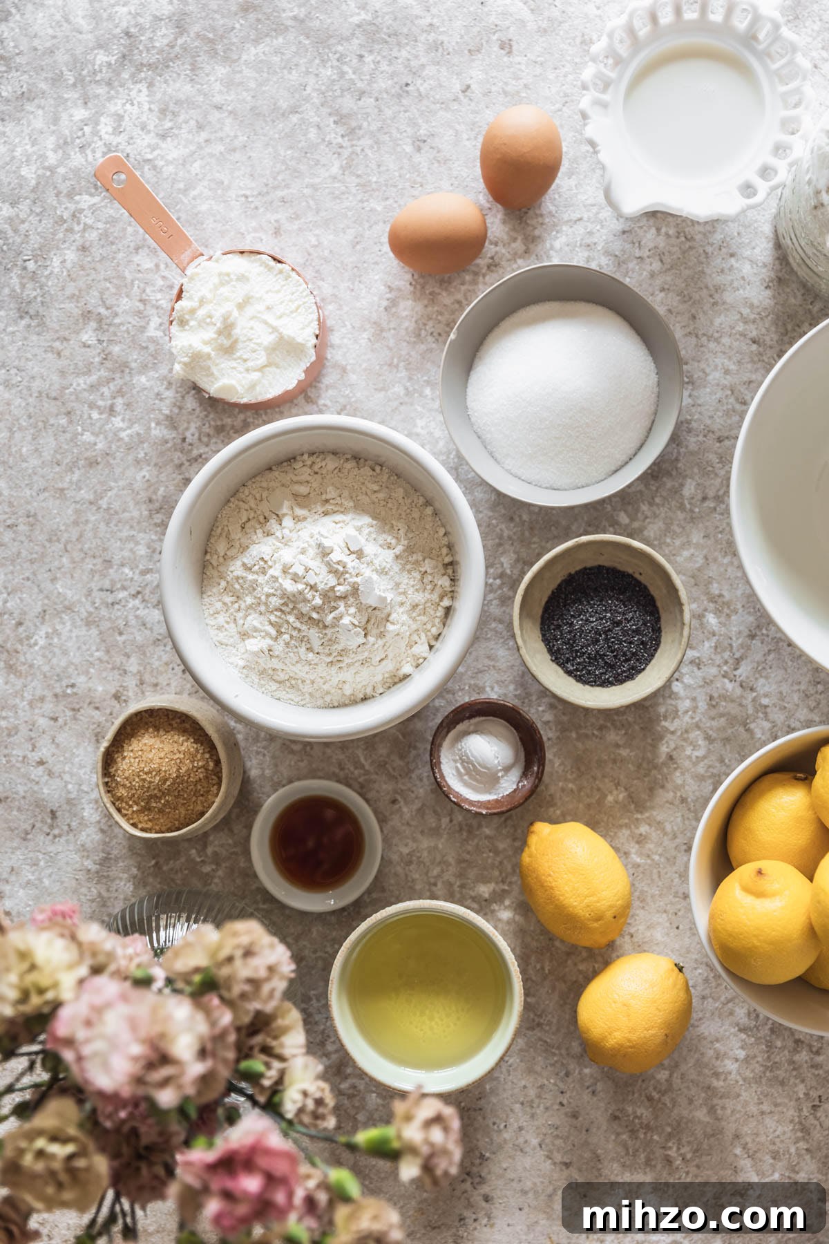 Ricotta Lemon Poppy Seed Muffins with Zesty Citrus Glaze 3 Various white and grey bowls with baking ingredients on a beige table next to a bouquet of pink flowers and white bowl of lemons.