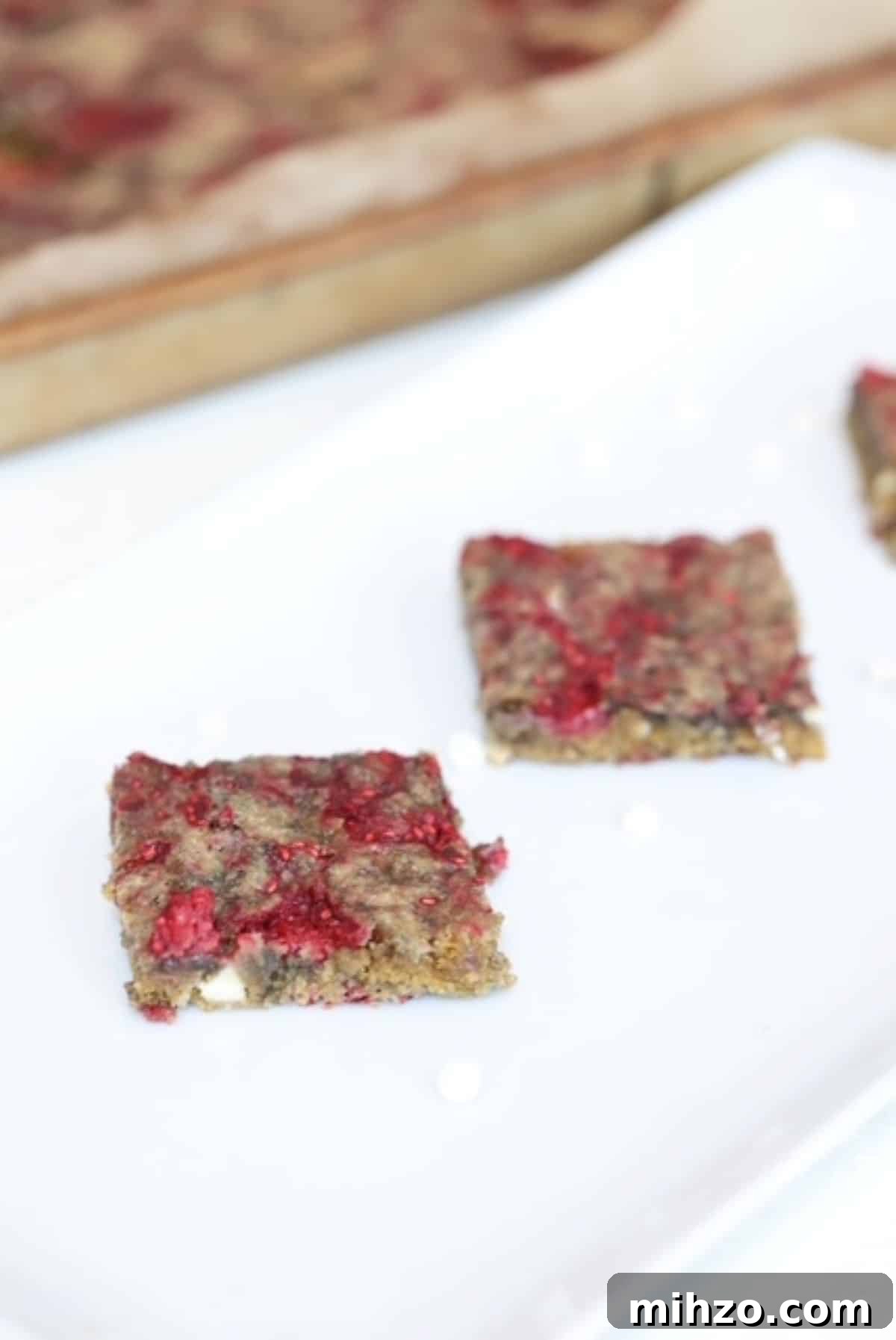 Raspberry White Chocolate Blondies with Raspberry Swirl 2 Two raspberry white chocolate blondies on a serving platter with a lined bar pan of blondies in the background.