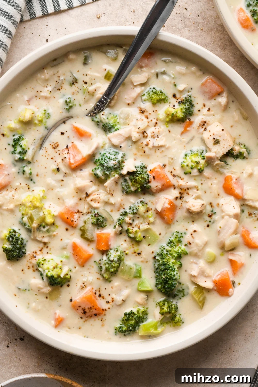 Delicious Dairy-Free Chicken and Broccoli Soup Recipe 19 A shallow cream colored ceramic bowl of creamy dairy free chicken and broccoli soup on a light brown background. A spoon is tucked into the edge of the bowl and a striped napkin is next to it.