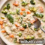 A shallow cream colored ceramic bowl of creamy dairy free chicken and broccoli soup on a light brown background. A spoon is digging into the middle of the soup for a bite, and a striped napkin is next to the bowl.