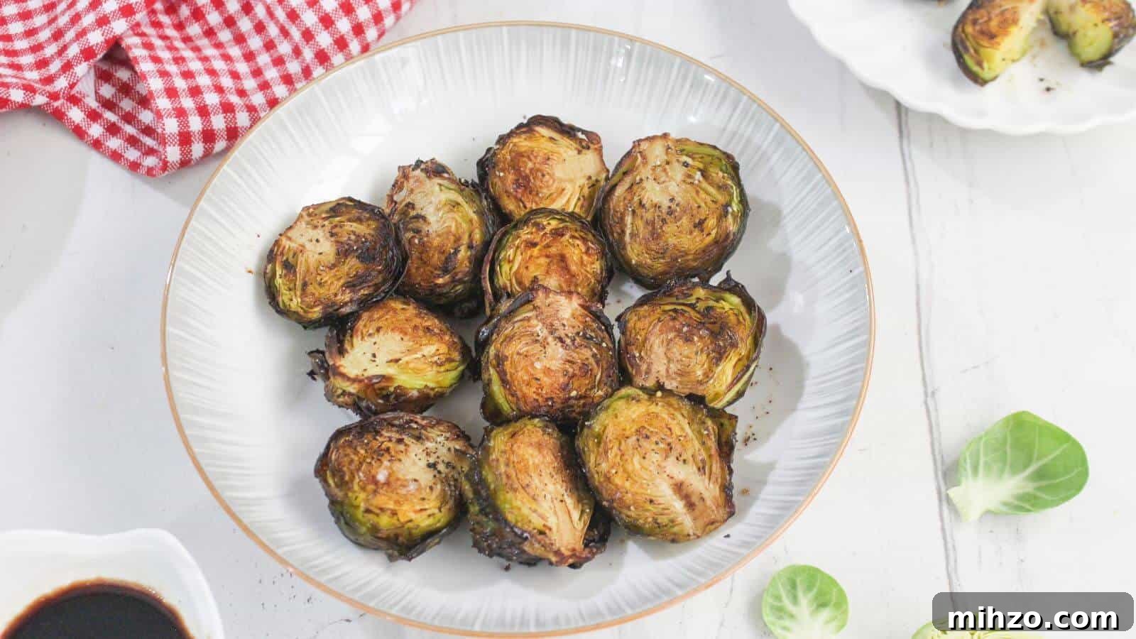 24 Brussels Sprouts Recipes That Even Non-Veggies Will Love 4 A white bowl filled with roasted Brussels sprouts, placed on a white surface with a red checkered cloth and scattered Brussels sprout leaves nearby.