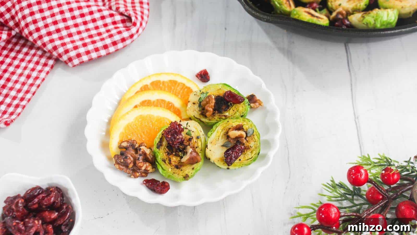 24 Brussels Sprouts Recipes That Even Non-Veggies Will Love 7 White plate with roasted Brussels sprouts halves, walnuts, dried cranberries, and three orange slices, set on a white surface near a red-checked cloth and holiday berries.