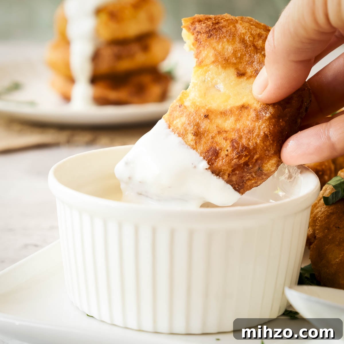 Crispy Potato Fritters Recipe — Golden Pan-Fried Latkes 9 Side shot of a hand dipping a potato fritter in cheese sauce in a ramekin with more fritters in the background.