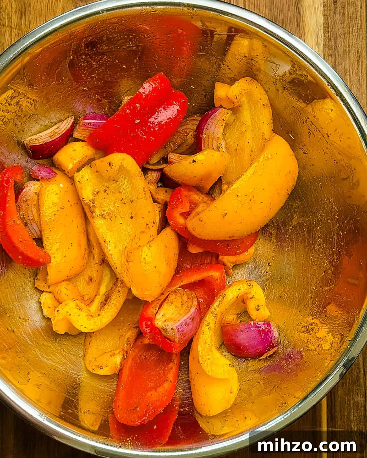 Sliced bell peppers and red onions tossed with olive oil and seasoning in a stainless steel mixing bowl.