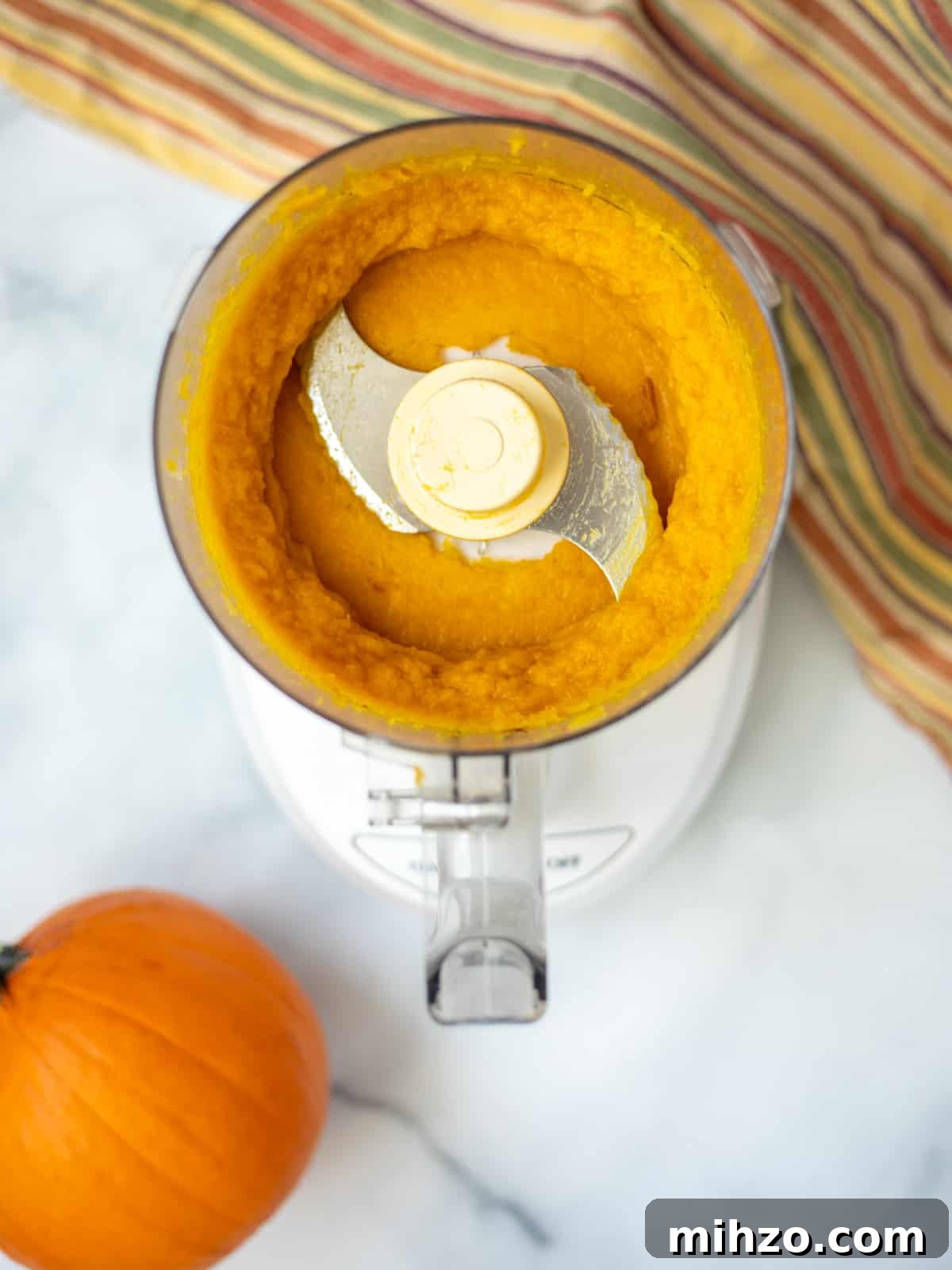 Food processor with pumpkin puree inside bowl with sugar pumpkin to side.