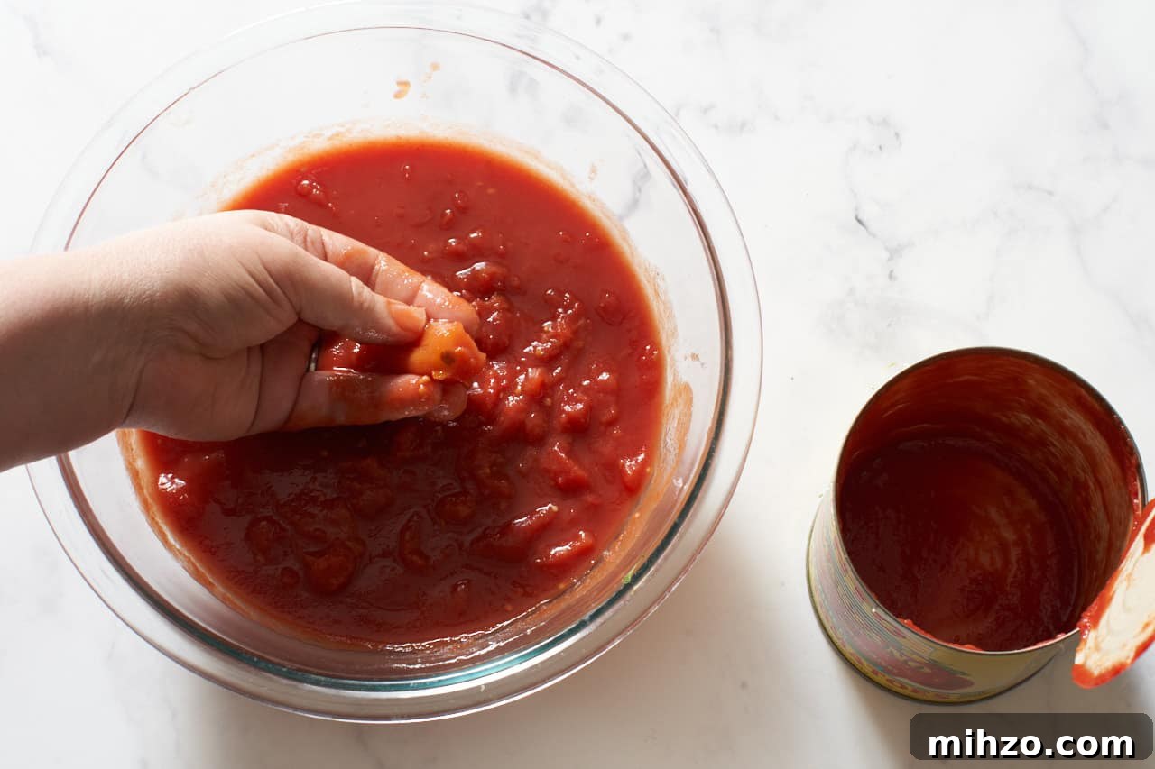 Authentic San Marzano Tomato Sauce Recipe for Rich Flavor 4 A woman's hand shown crushing San Marzano tomatoes that have been poured into a bowl. An empty can is on the right.
