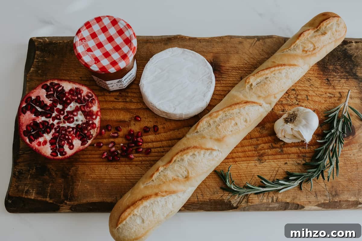 rectangular wooden board topped with a jar of bonne maman apricot preserves, a halved pomegranate, a round of brie, a baguette, a head of garlic and a sprig of fresh rosemary.