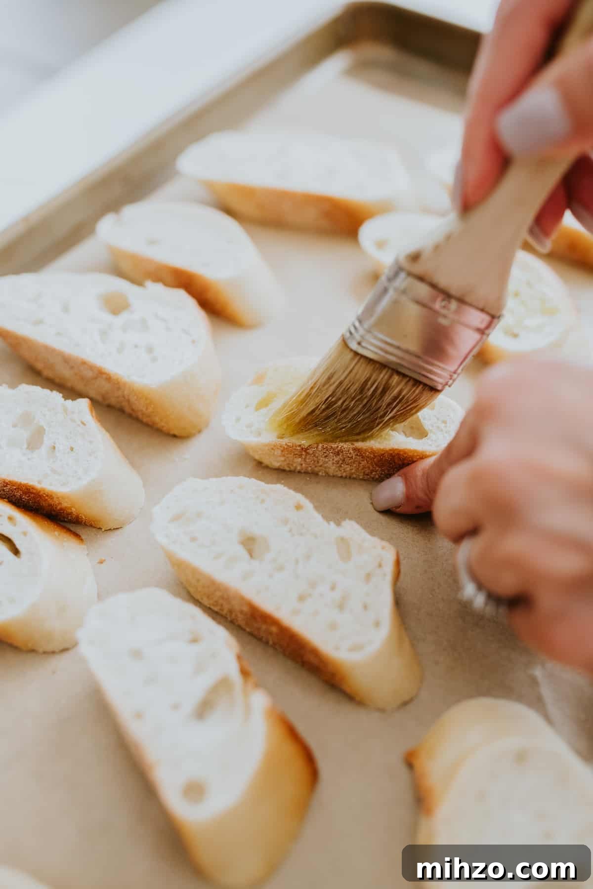 hands using a pastry brush to brush oil on a piece of sliced baguette on a parchment lined sheet tray.