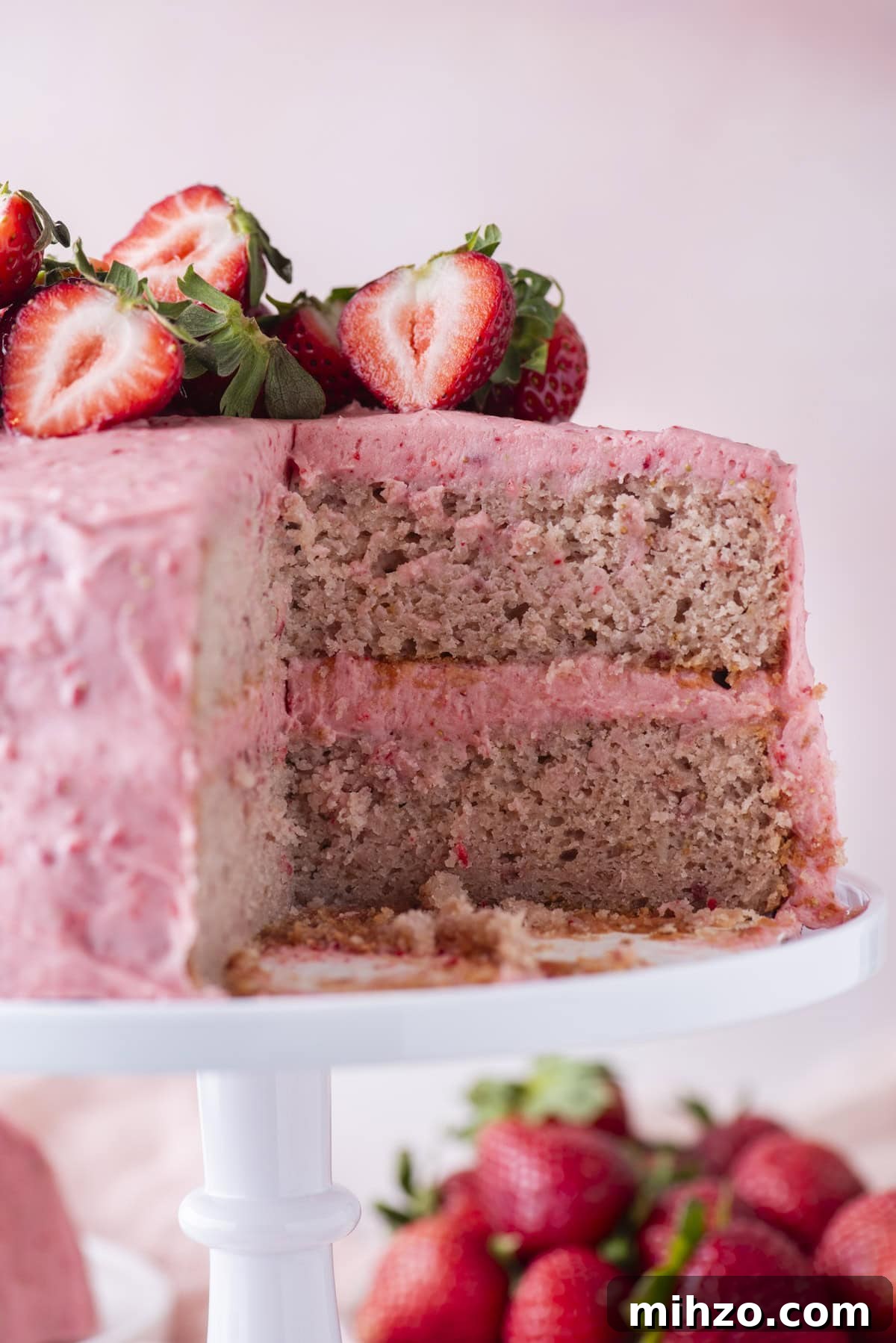 A two-layer strawberry cake with pink frosting and fresh strawberries on top sits on a white cake stand. A large slice has been cut out, revealing the cake’s moist interior.