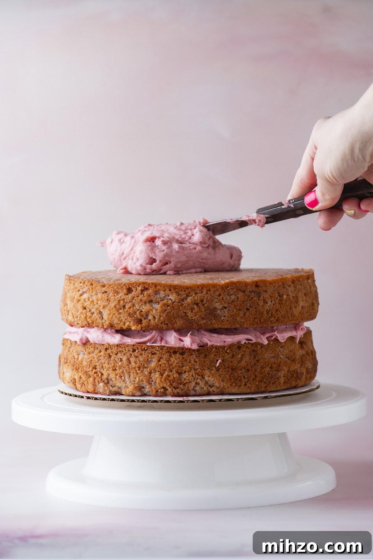 A hand spreads pink frosting on top of a two-layer cake with pink filling, placed on a white cake stand against a soft pink background.