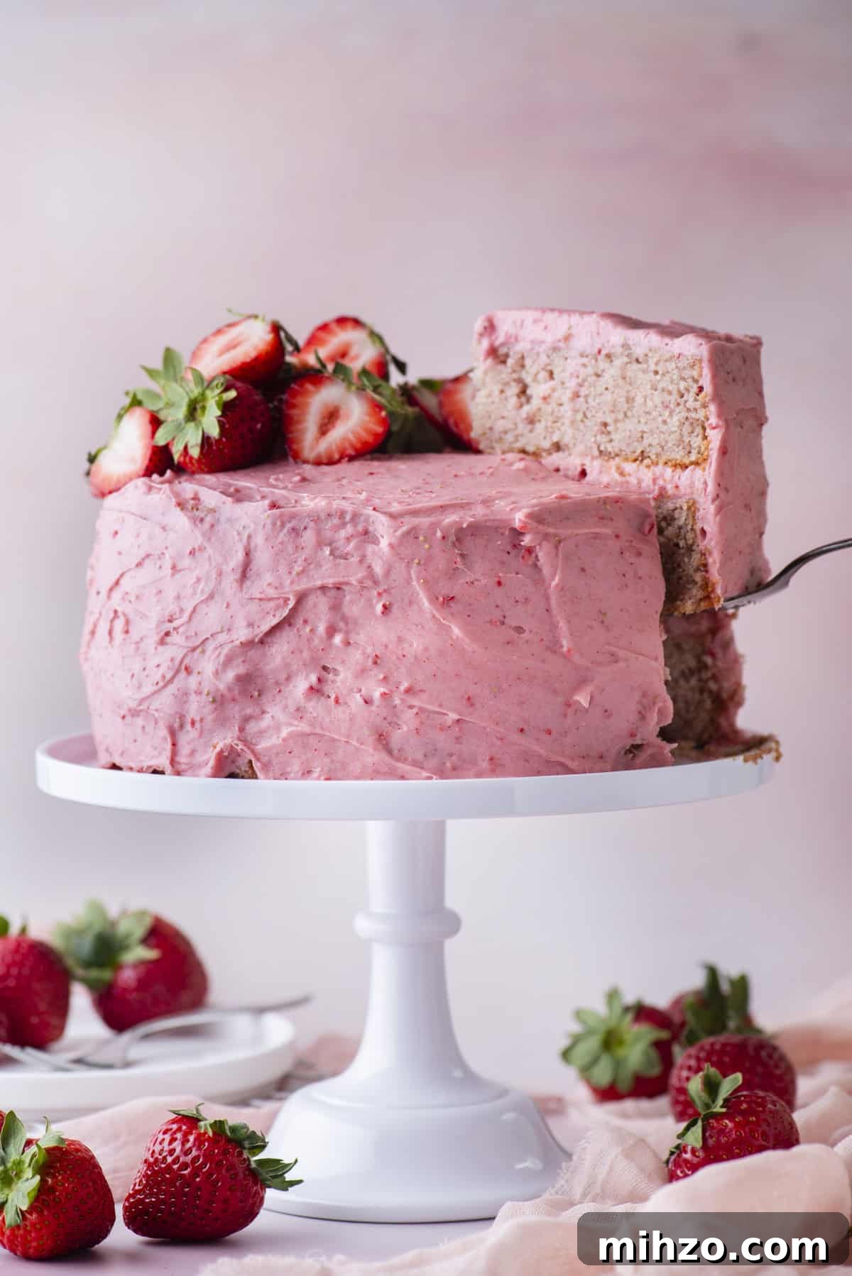 A strawberry cake with pink frosting and fresh strawberries on top sits on a white cake stand. A slice is being lifted out, revealing the cake layers inside. Strawberries are scattered around the base.