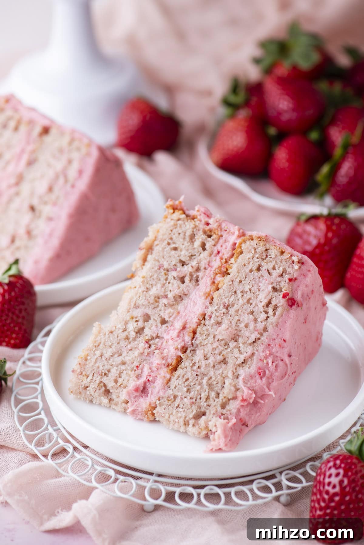 A slice of strawberry cake with pink frosting sits on a white plate, with fresh strawberries scattered around and a second slice of cake in the background.