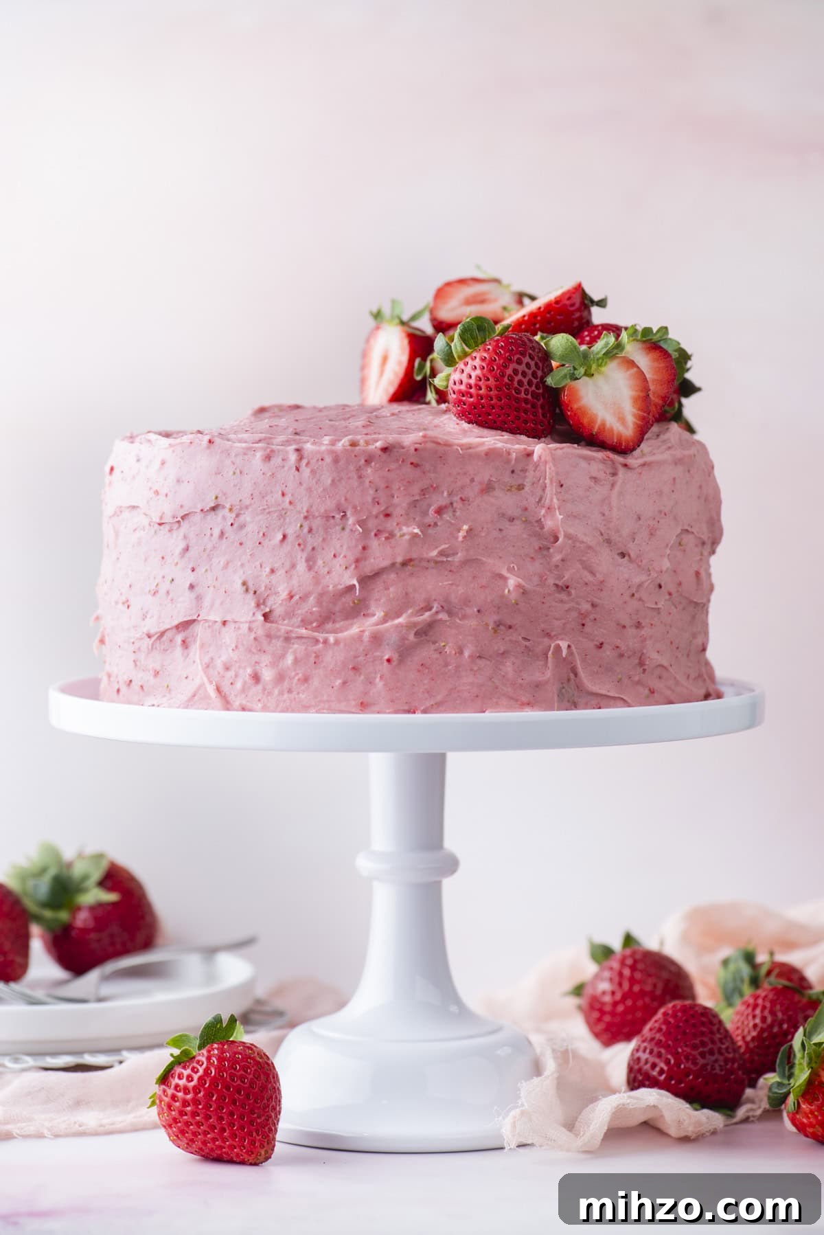 A round cake with pink strawberry frosting sits on a white cake stand, topped with fresh strawberries. Whole and halved strawberries are scattered around the base, with a soft pink background and plates in the distance.