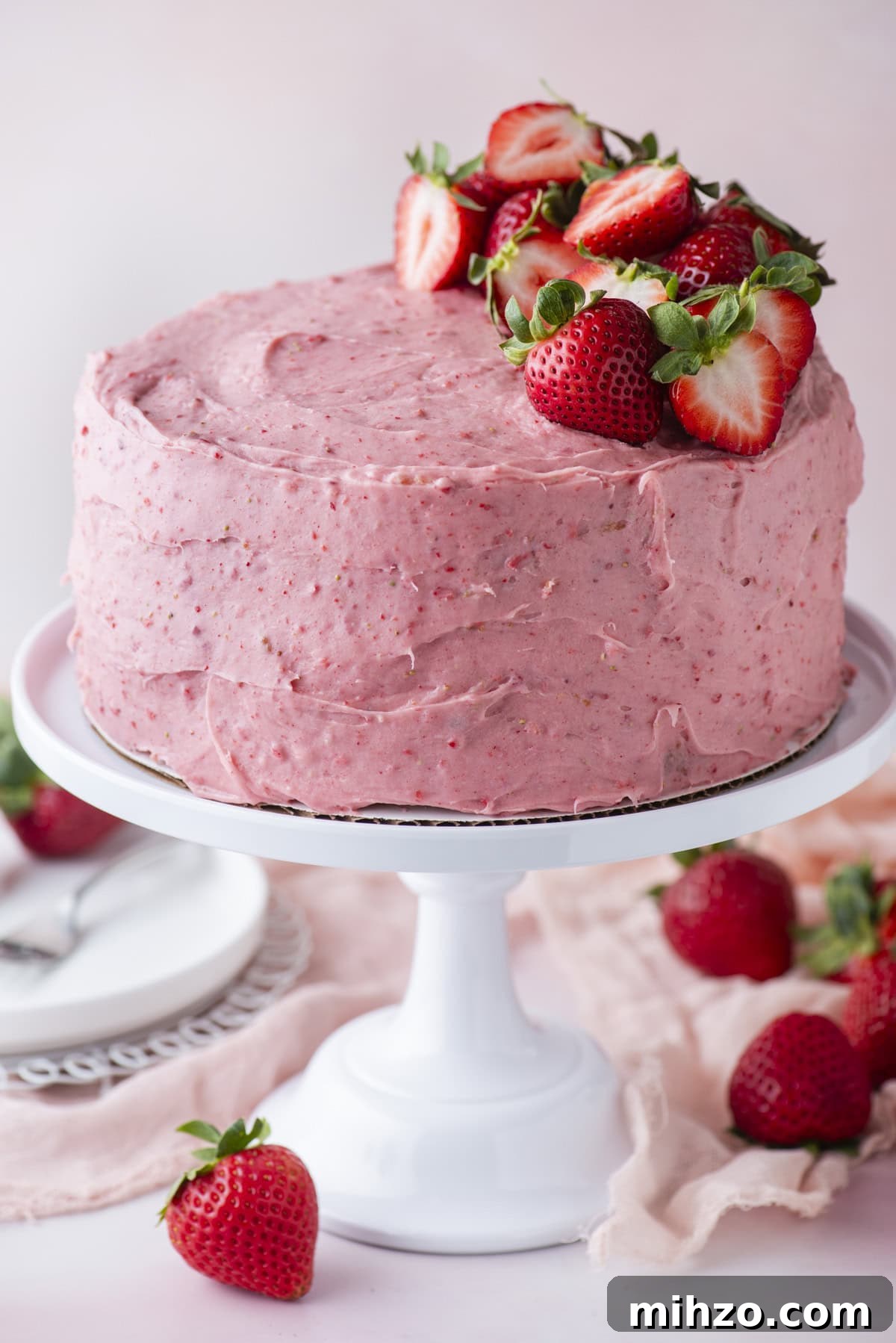 A round cake with pink strawberry frosting sits on a white cake stand, topped with fresh strawberries. Whole strawberries are scattered around the base, and plates and utensils are visible in the background.