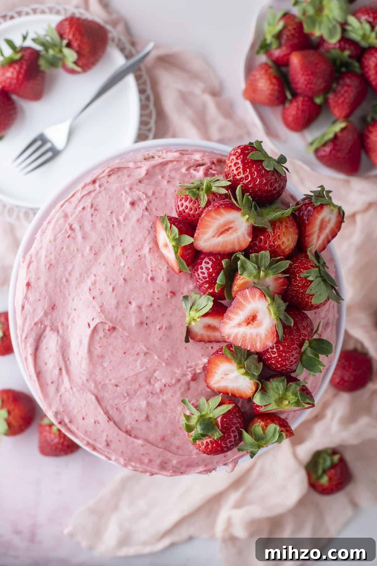 A cake topped with pink strawberry frosting and fresh strawberries, some halved, sits on a light pink surface with whole strawberries and a plate with a fork nearby.