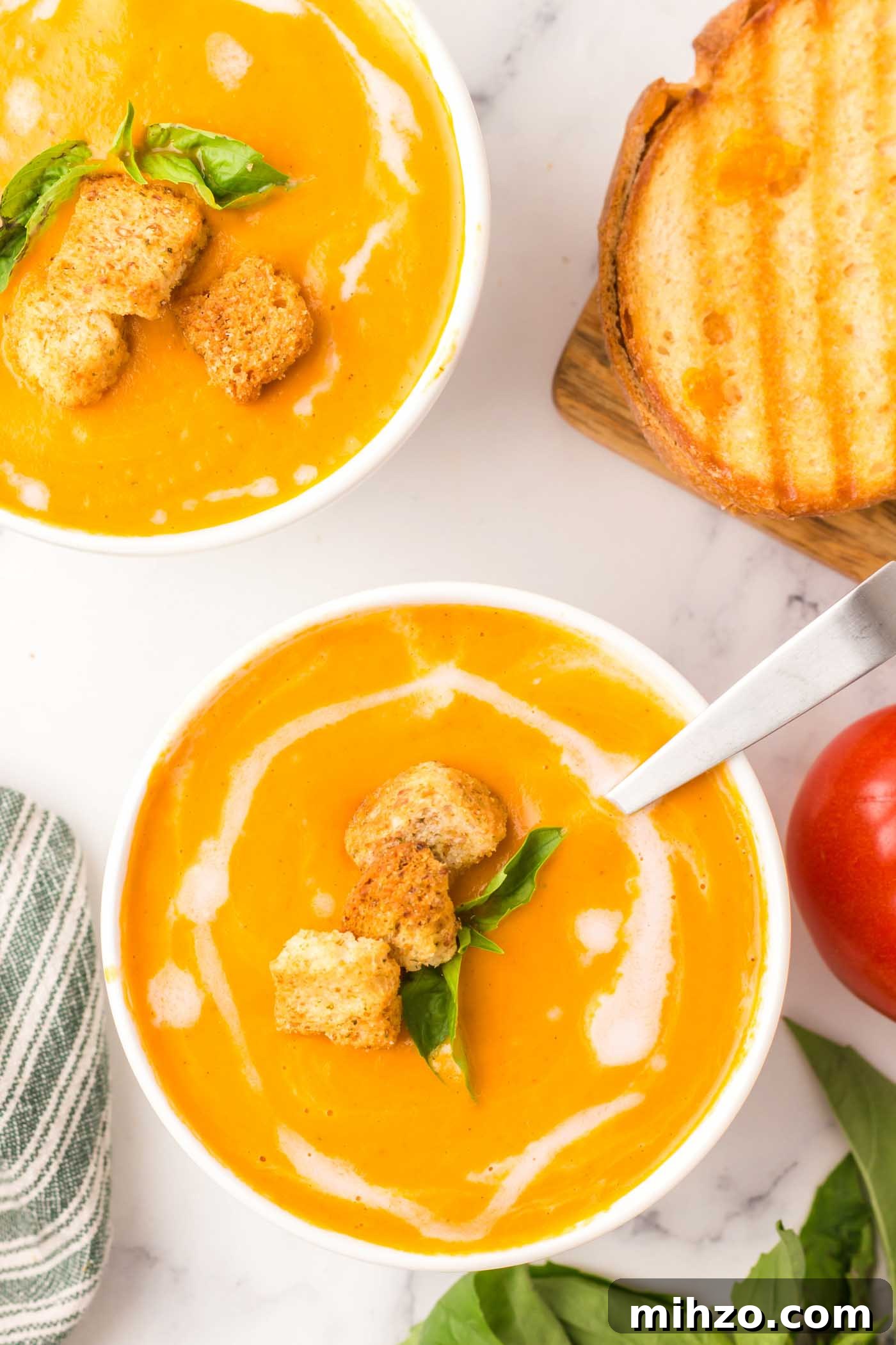 Two bowls of creamy vegan tomato soup topped with a drizzle of coconut oil and two croutons with a few slices of toasted bread beside them.