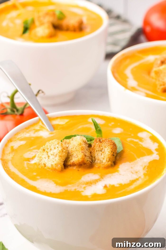 A bowl of creamy tomato soup drizzled with cream and topped with croutons and a piece of fresh basil.