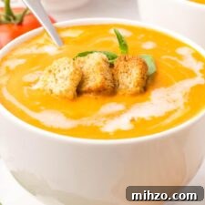 A bowl of creamy tomato soup drizzled with cream and topped with croutons and a piece of fresh basil.