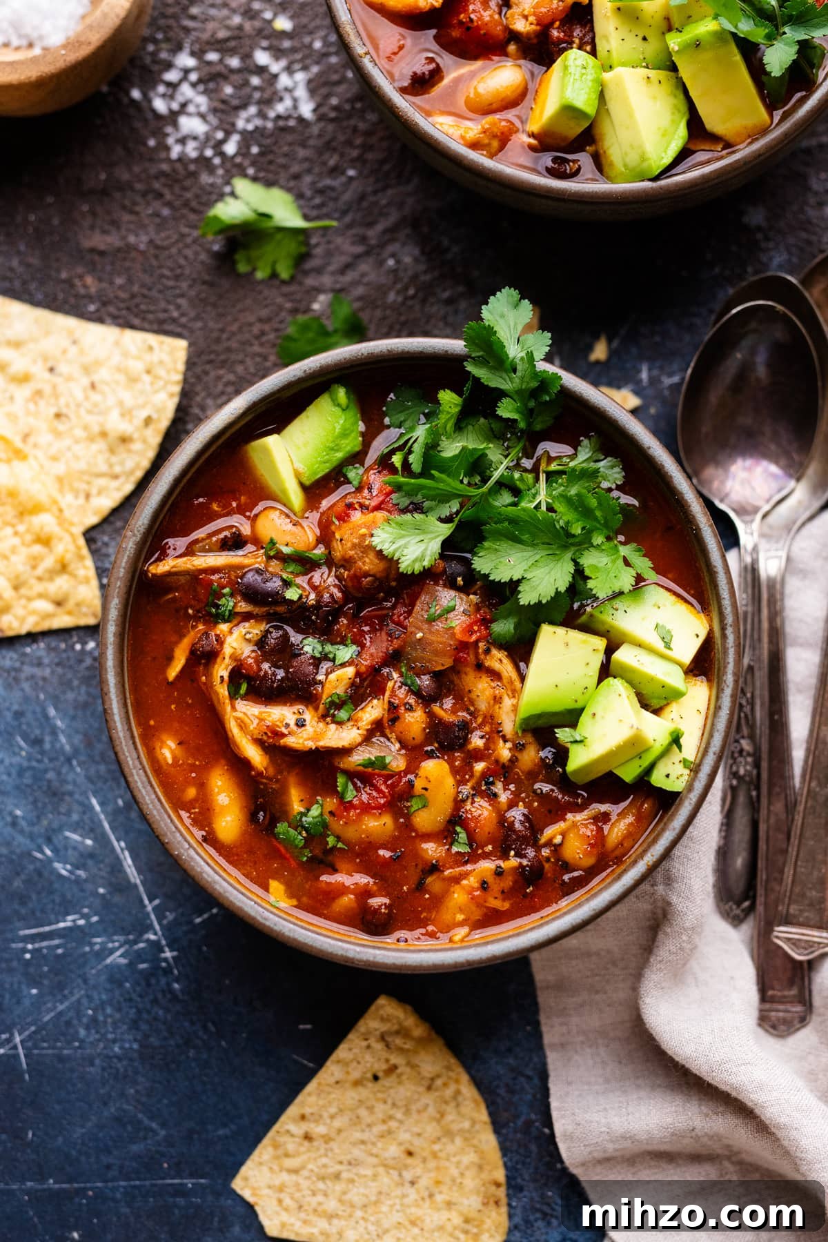 Slow-Cooker Chicken Chili Recipe for Cozy Weeknight Meals 2 A gray bowl with red chicken chili in it. The bowl of chili is sitting on a dark gray background with tortilla chips, green herbs, and a napkin with spoons arranged around it.