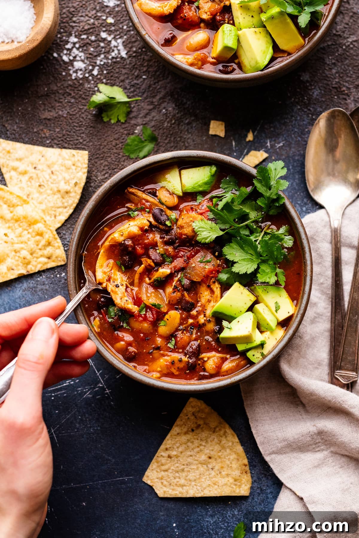 Slow-Cooker Chicken Chili Recipe for Cozy Weeknight Meals 6 A hand holding a spoon in a gray bowl with red chicken chili in it. The bowl of chili is arranged on a dark gray background with tortilla chips, cilantro herbs, and a napkin with spoons on it.