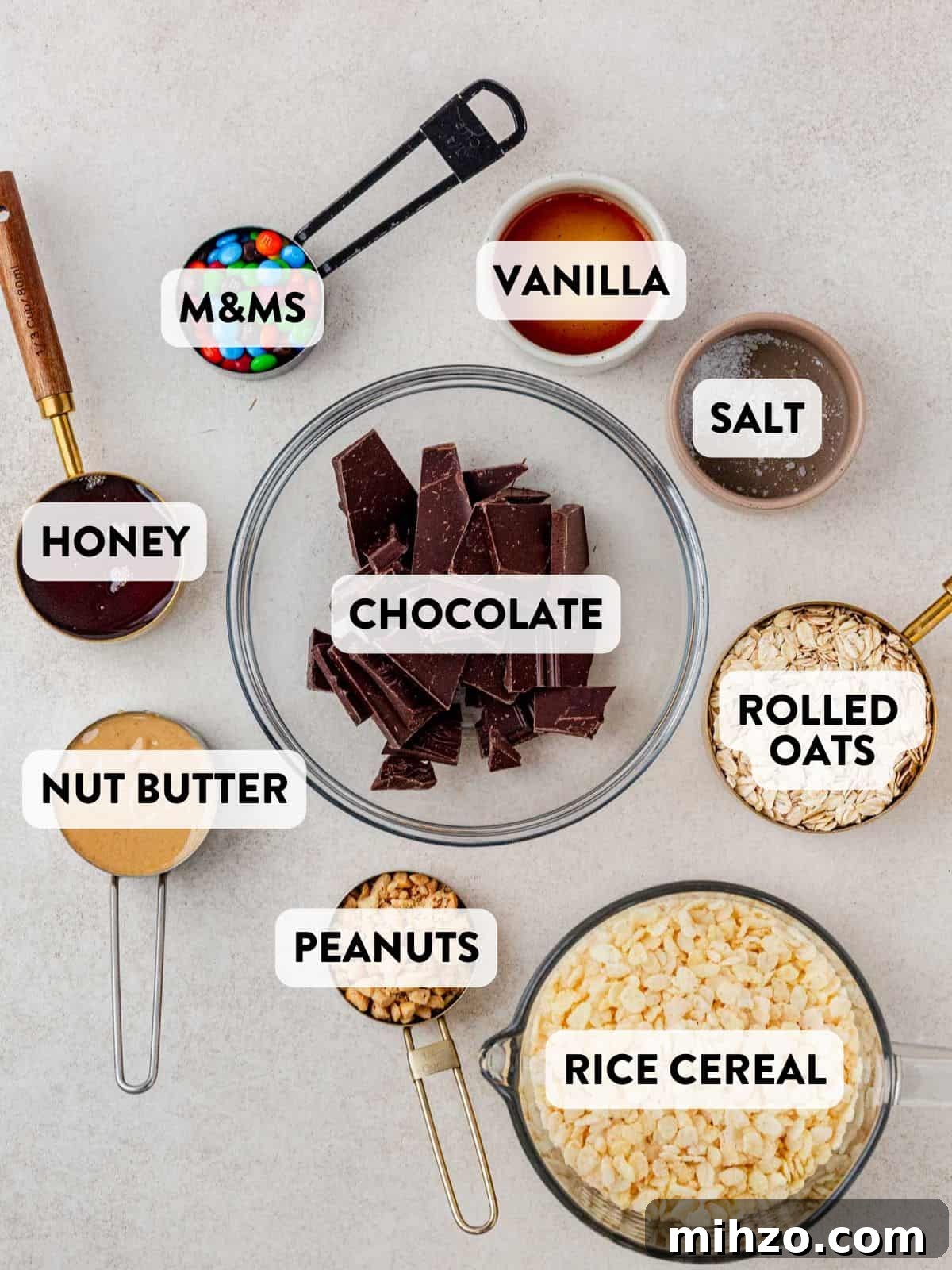 Flat lay of homemade Kudos granola bar ingredients on a light background, including chocolate chunks, M&Ms, vanilla extract, salt, honey, nut butter, peanuts, rolled oats, and rice cereal—all labeled for reference.
