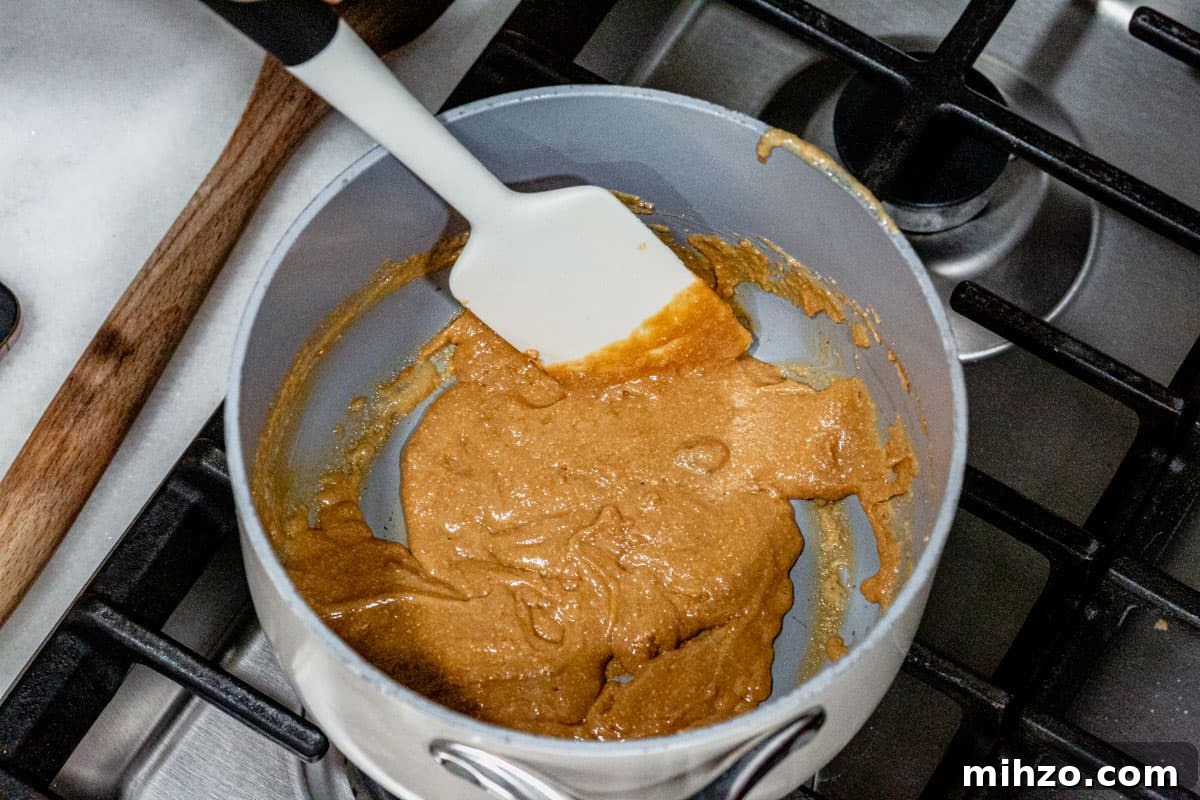 Nut butter and honey mixture bubbling and being stirred with a spatula in a white saucepan over a gas stovetop.