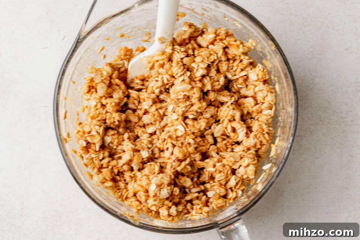 Rolled oats and rice cereal mixture coated in nut butter and honey, stirred together in a glass mixing bowl with a white spatula.