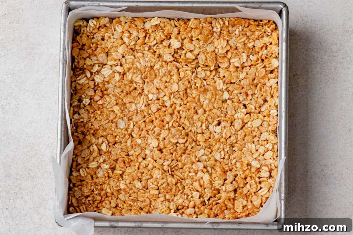 Granola bar mixture pressed firmly into a parchment-lined square baking pan, ready to chill in the fridge.
