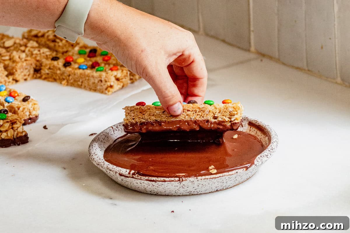 Hand dipping a homemade copycat Kudos granola bar topped with mini M&Ms into melted chocolate for a fun and easy no-bake treat.