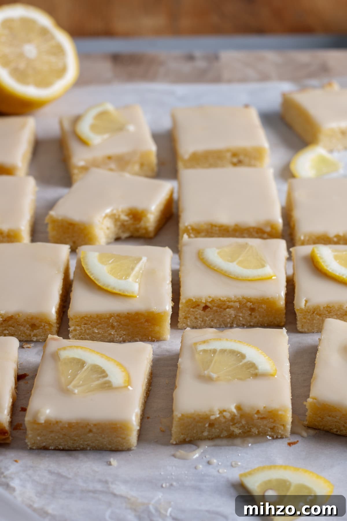 Zesty Lemon Blondies with Luscious Glaze 15 Lemon blondies topped with thin slices of lemon on a piece of parchment paper.