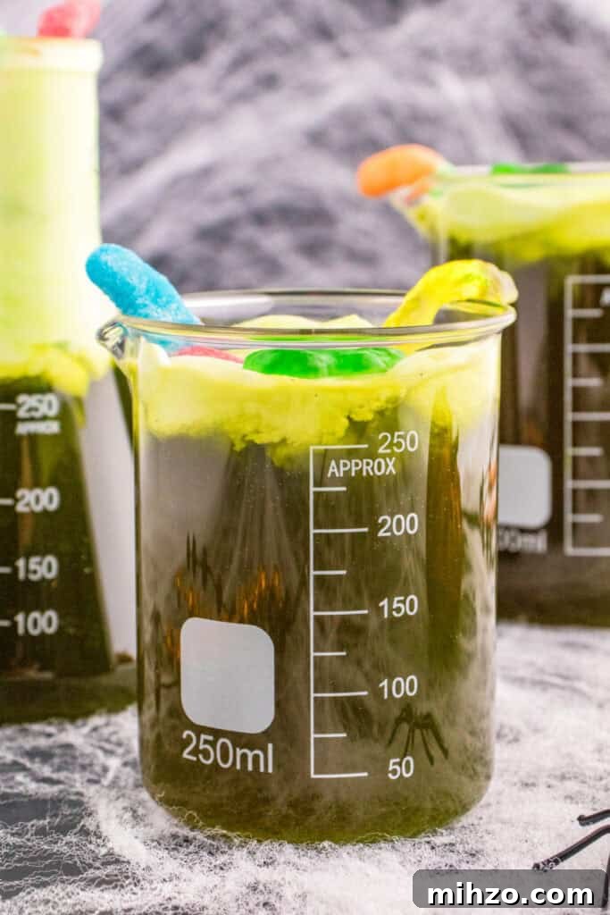 Toxic Waste Punch Recipe: How to Make a Neon Party Drink 2 Close-up of a beaker filled with Toxic Waste Punch, topped with green foam and gummy decorations, with cobwebs in the background.