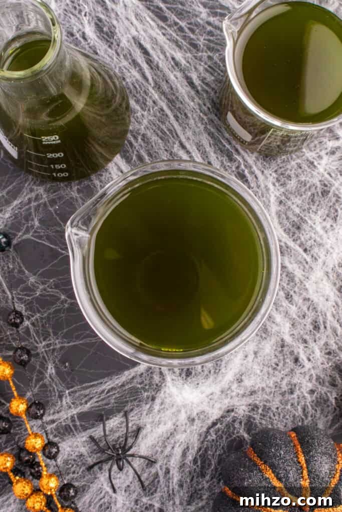Toxic Waste Punch Recipe: How to Make a Neon Party Drink 14 Three glass beakers filled with dark green Toxic Waste Punch on a cobweb-decorated table with Halloween beads and pumpkins.