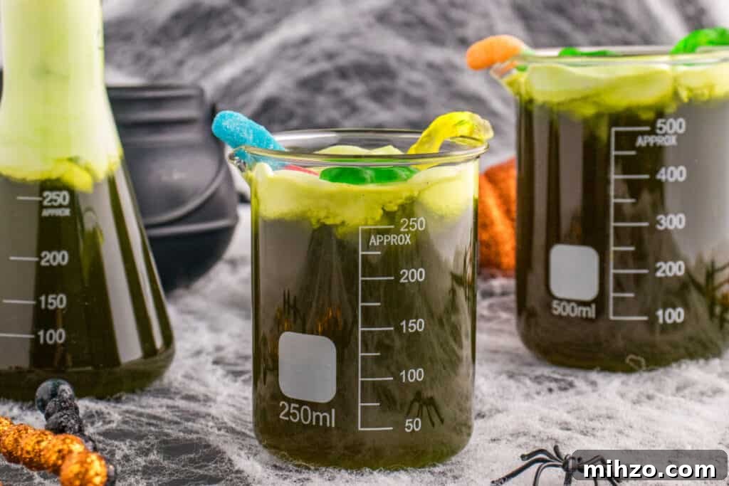 Toxic Waste Punch Recipe: How to Make a Neon Party Drink 18 Halloween table with multiple beakers of Toxic Waste Punch topped with green foam and gummy candy, surrounded by cobwebs and spiders.