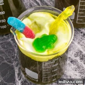 Toxic Waste Punch Recipe: How to Make a Neon Party Drink 23 Square cropped image of a beaker filled with Toxic Waste Punch topped with foamy green whipped cream and gummy candy decorations.