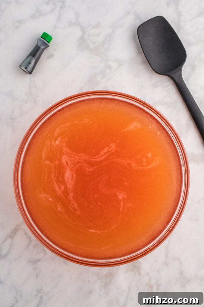 Toxic Waste Punch Recipe: How to Make a Neon Party Drink 6 Large glass bowl filled with orange punch mixture and a black spatula on the side with green food coloring bottle nearby.
